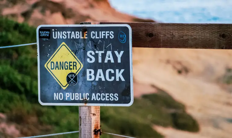 Warning sign indicating danger due to unstable cliffs, advises staying back. Outdoors setting.